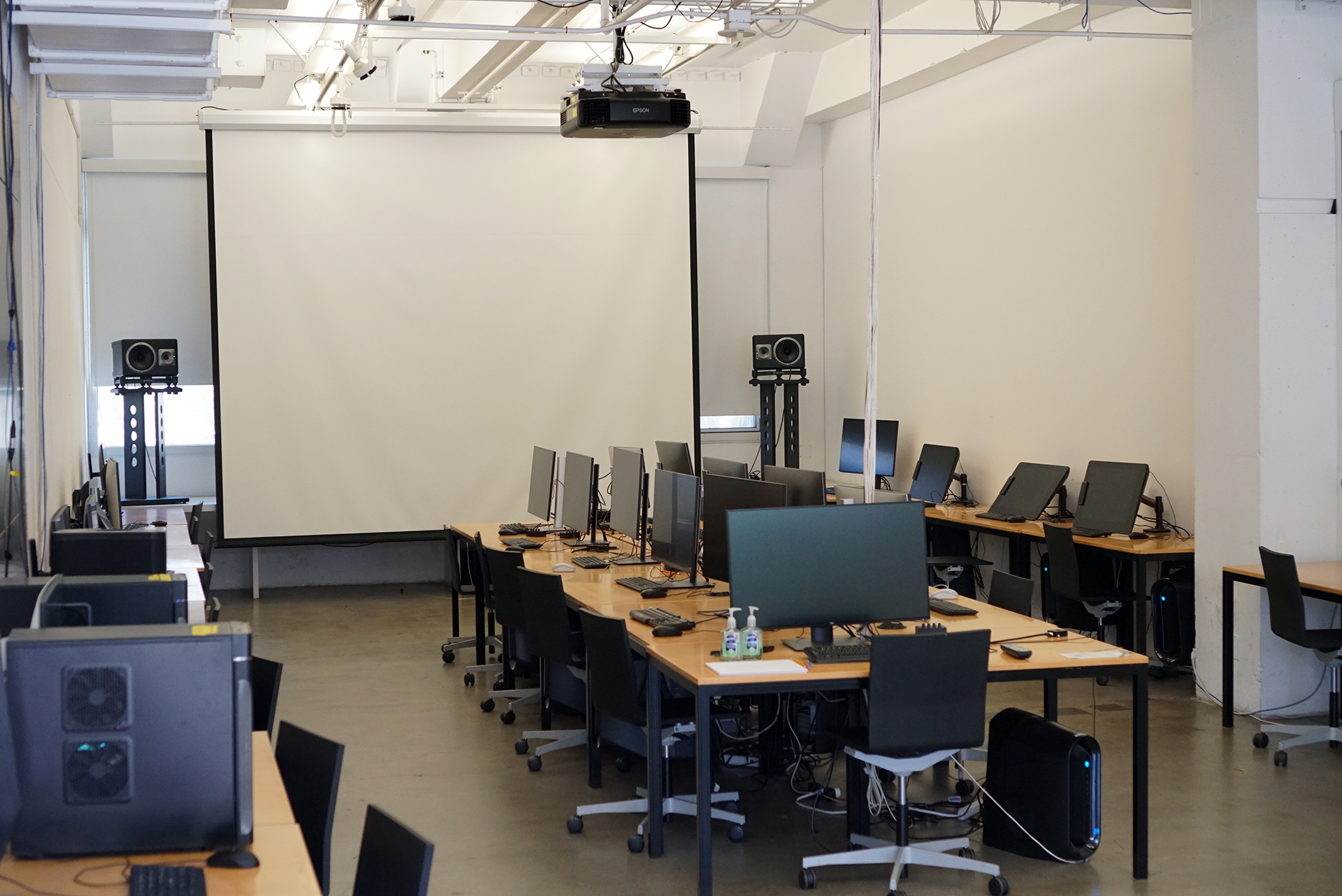 A wide shot of the 3D Classroom, displaying a central arrangement of wooden desks equipped with monitors and black rolling chairs, all oriented toward a large projector screen at the front of the room.
