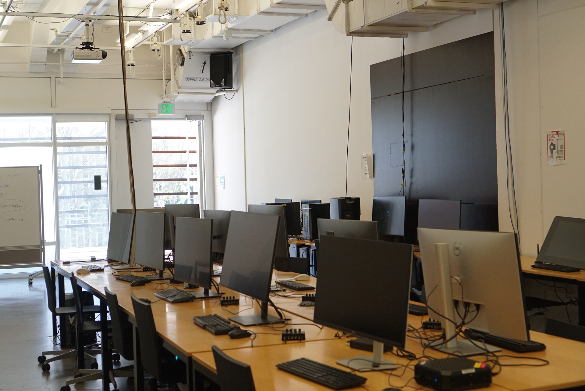 An angled view from the back of the 3D Classroom, featuring rows of monitors on wooden desks. A large black panel is mounted on the right wall, with an exit door and bright windows visible in the background.