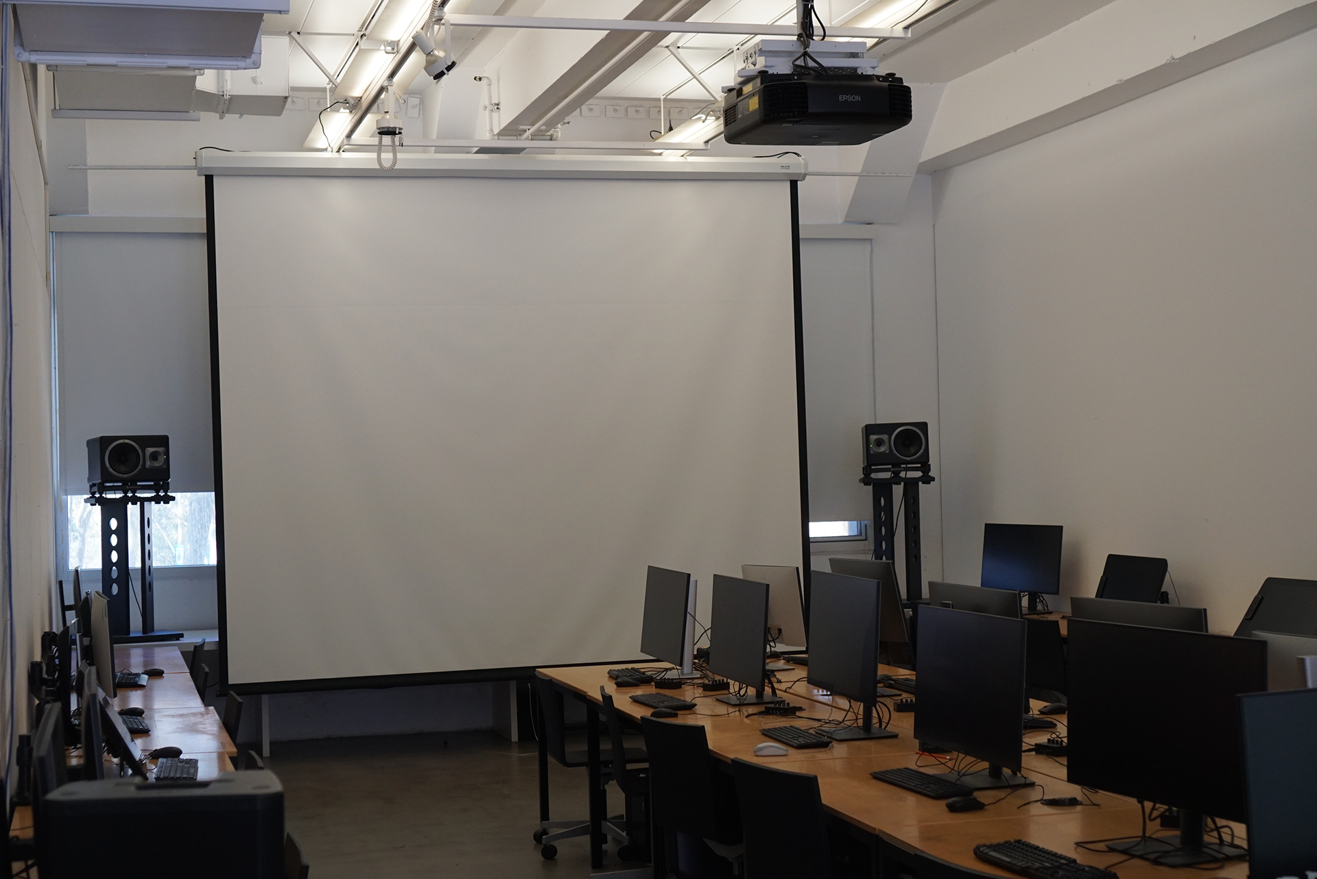 A view of the 3D Classroom showing rows of computers on wooden desks facing a large, lowered projector screen. Speakers on black stands flank the screen, and a projector is mounted to the exposed white ceiling.