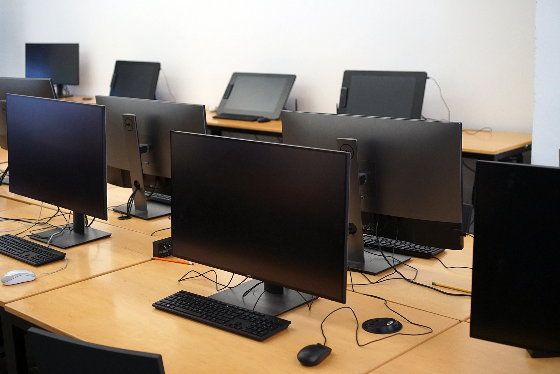 A close-up view of wooden computer desks in the 3D Classroom. Several large monitors and keyboards are visible in the foreground, while digital drawing tablets are set up on the desks in the background against a white wall.