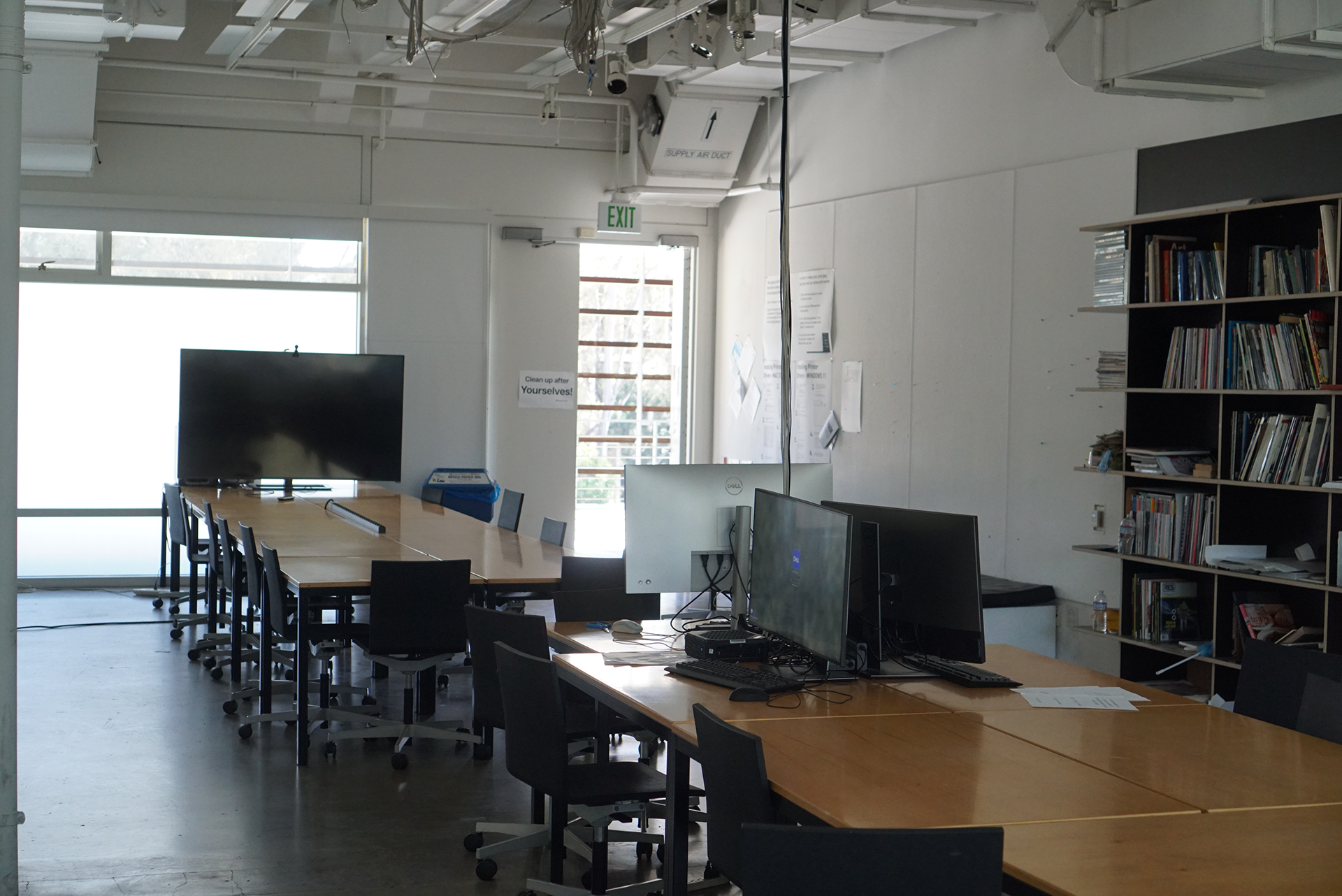 A wide shot of the Design Classroom displaying a long wooden table facing a large flat-screen monitor on the left, alongside additional computer workstations on wooden desks and a tall bookshelf against the right wall.