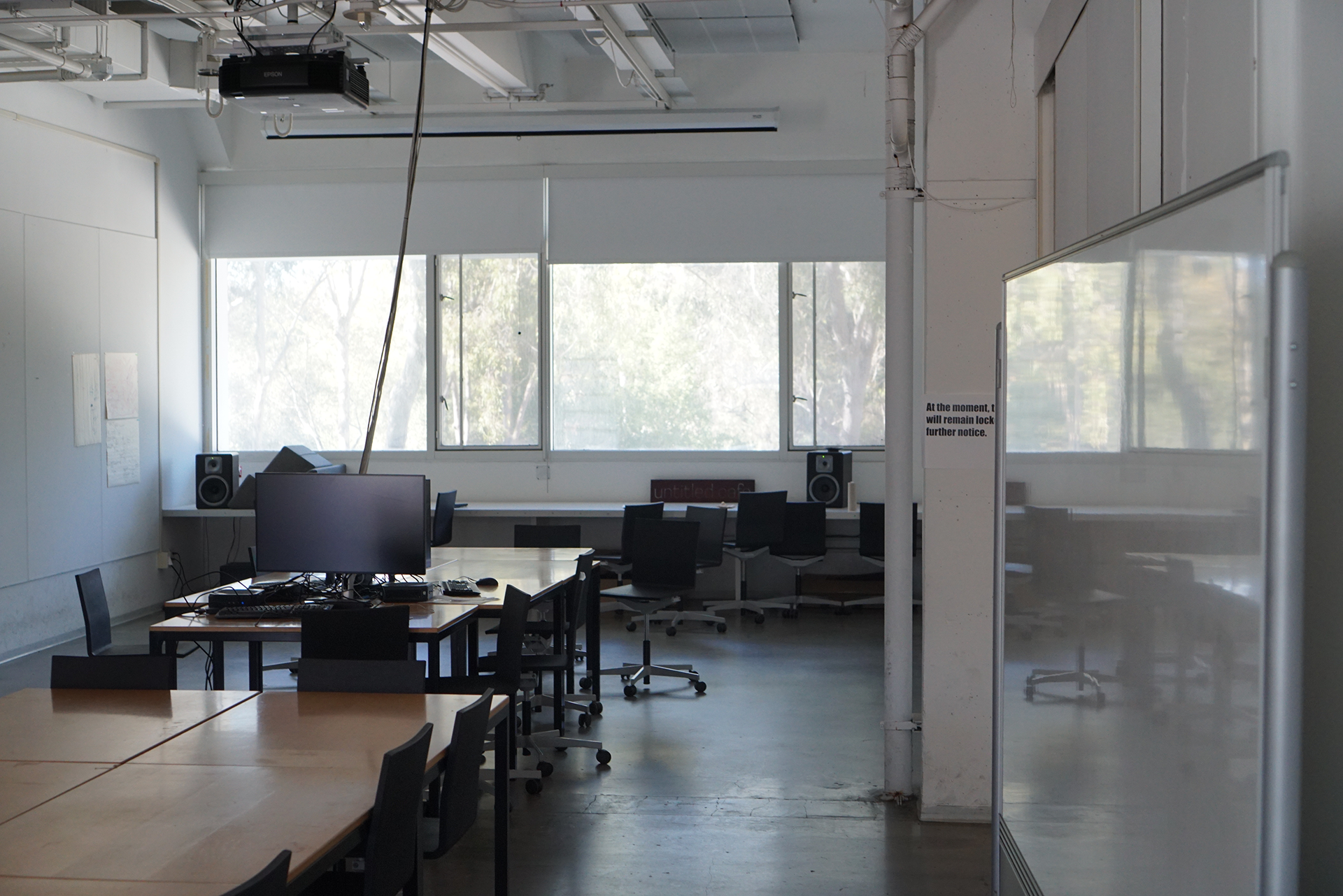An angled shot of the Design Classroom highlighting wooden desks equipped with a computer monitor, black rolling chairs, bright windows, a central white structural column, and a portable whiteboard on the right.