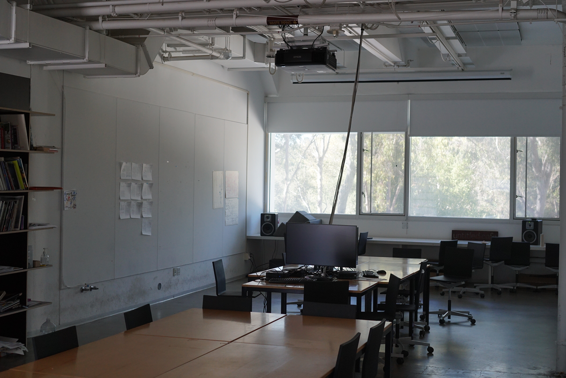 A view of the Design Classroom showing wooden tables and black rolling chairs arranged for collaboration, featuring a large whiteboard wall on the left, a ceiling-mounted projector, and wide windows looking out to trees.