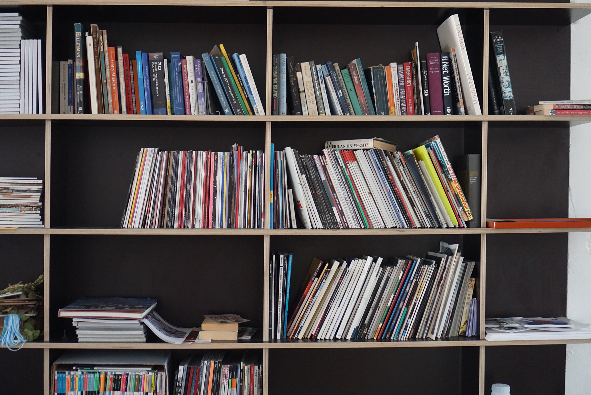 A close-up view of a tall, black shelving unit with light wood edges, partially filled with an assortment of colorful books, magazines, and reference materials.