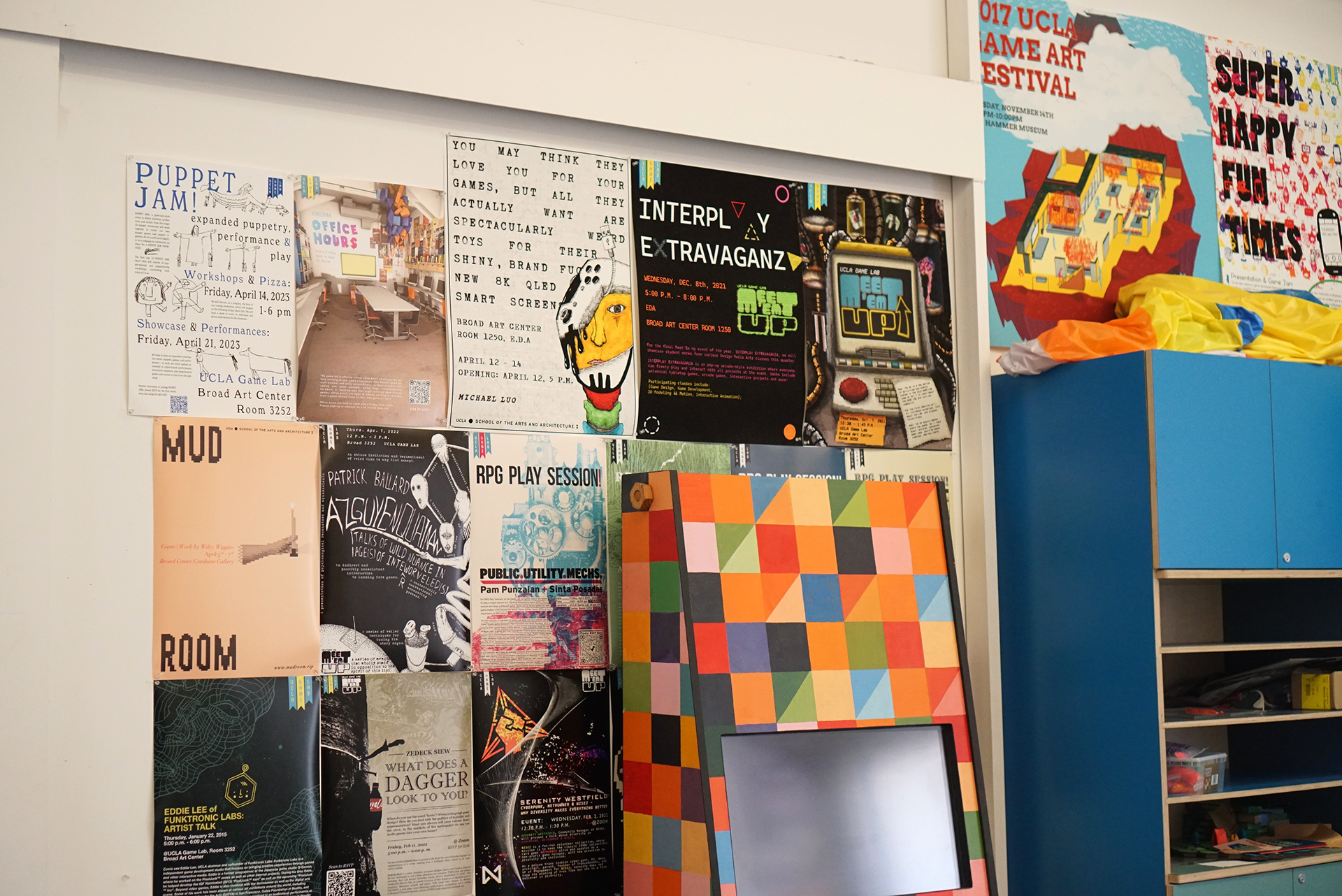 A section of a white wall densely covered with various colorful event posters, located next to a blue storage cabinet and a leaning frame displaying a multicolored checkerboard pattern.