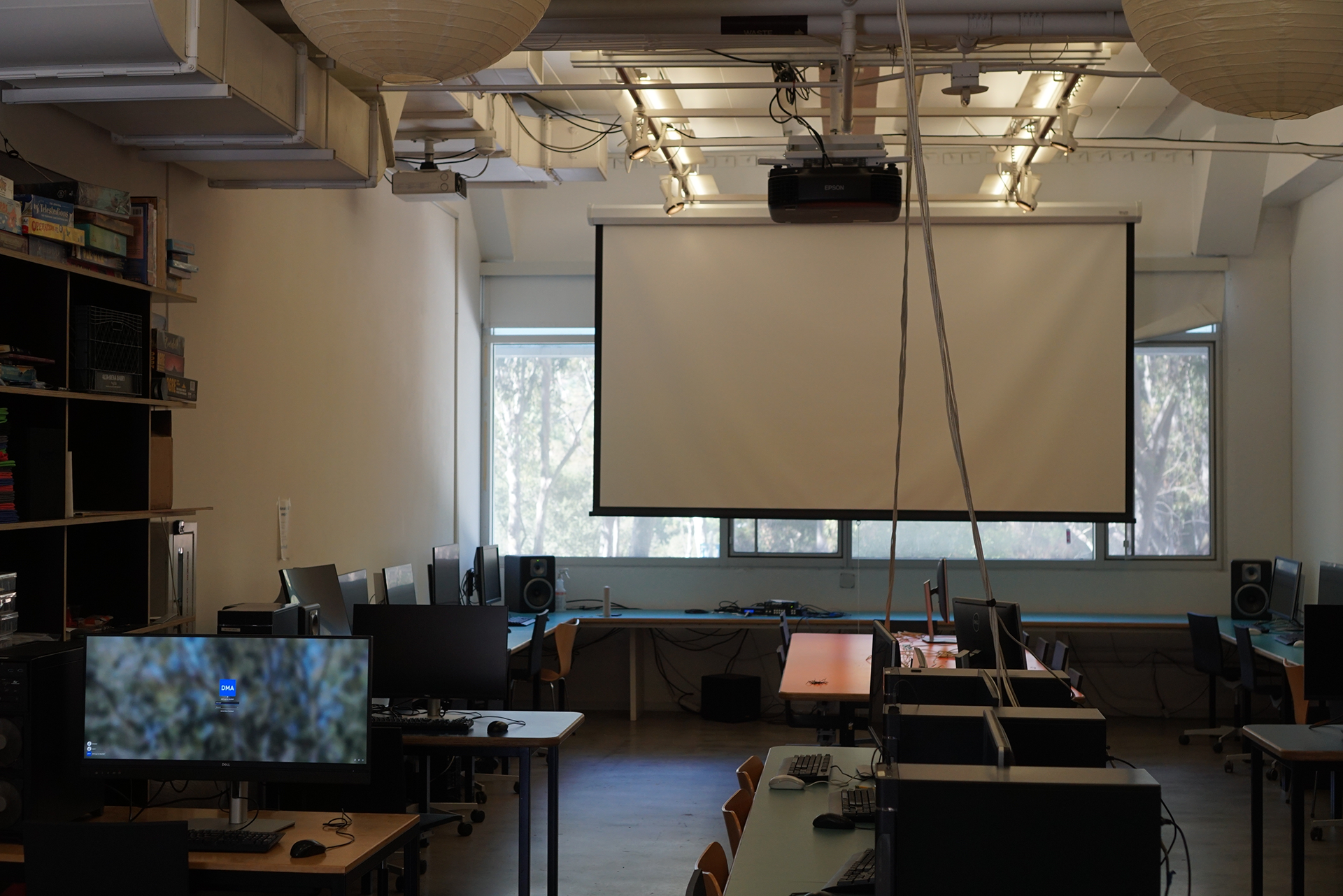 A straight-on view of the Games Classroom, showing a lowered projector screen hanging in front of a wide window, flanked by computer workstations on teal desks, with large round paper lanterns overhead and a tall shelf of board games visible on the left wall.