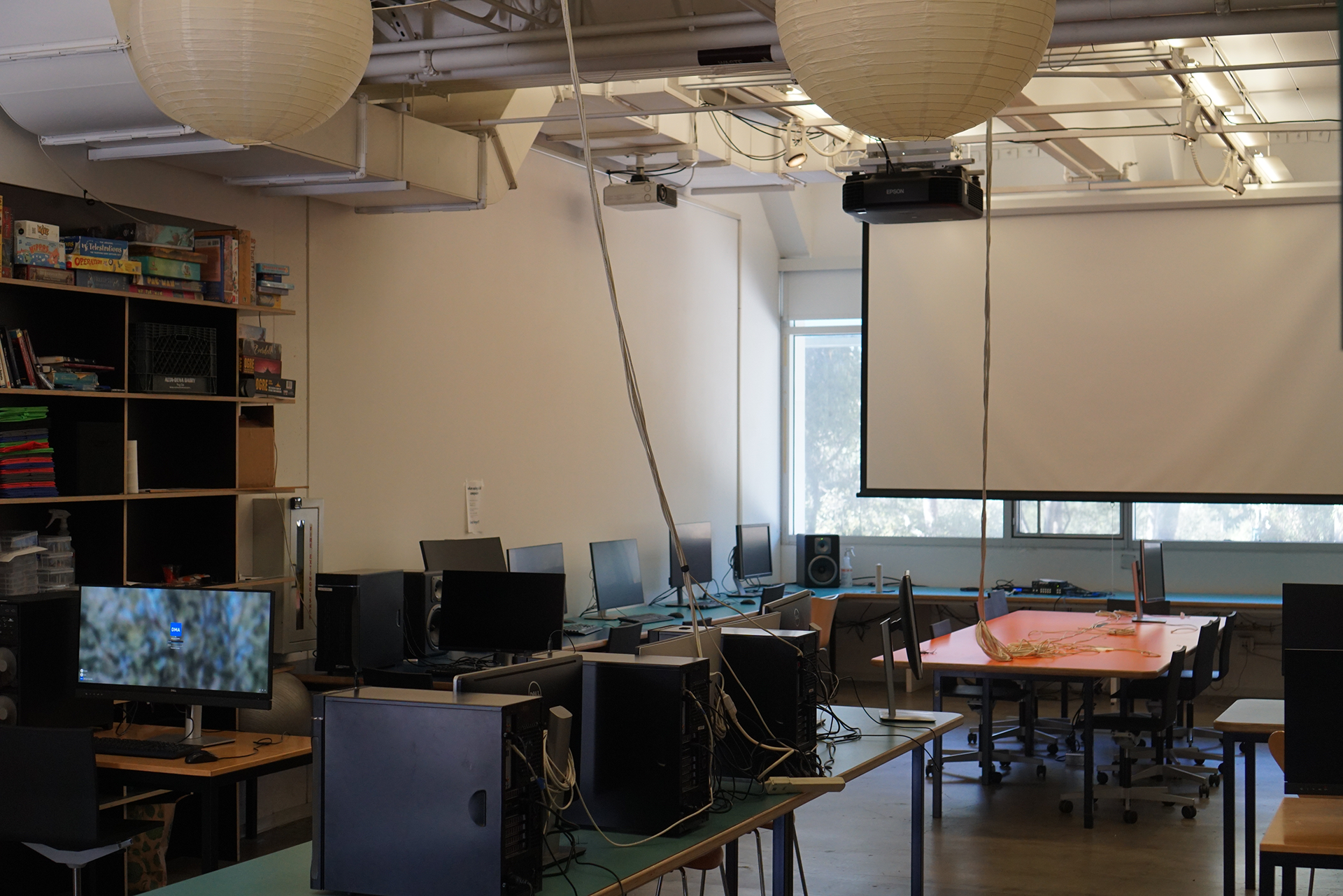 An angled view of the Games Classroom, featuring rows of computers on teal-topped desks, large spherical paper lanterns hanging from the ceiling, a lowered projector screen, and a tall wooden bookshelf filled with board games on the left.