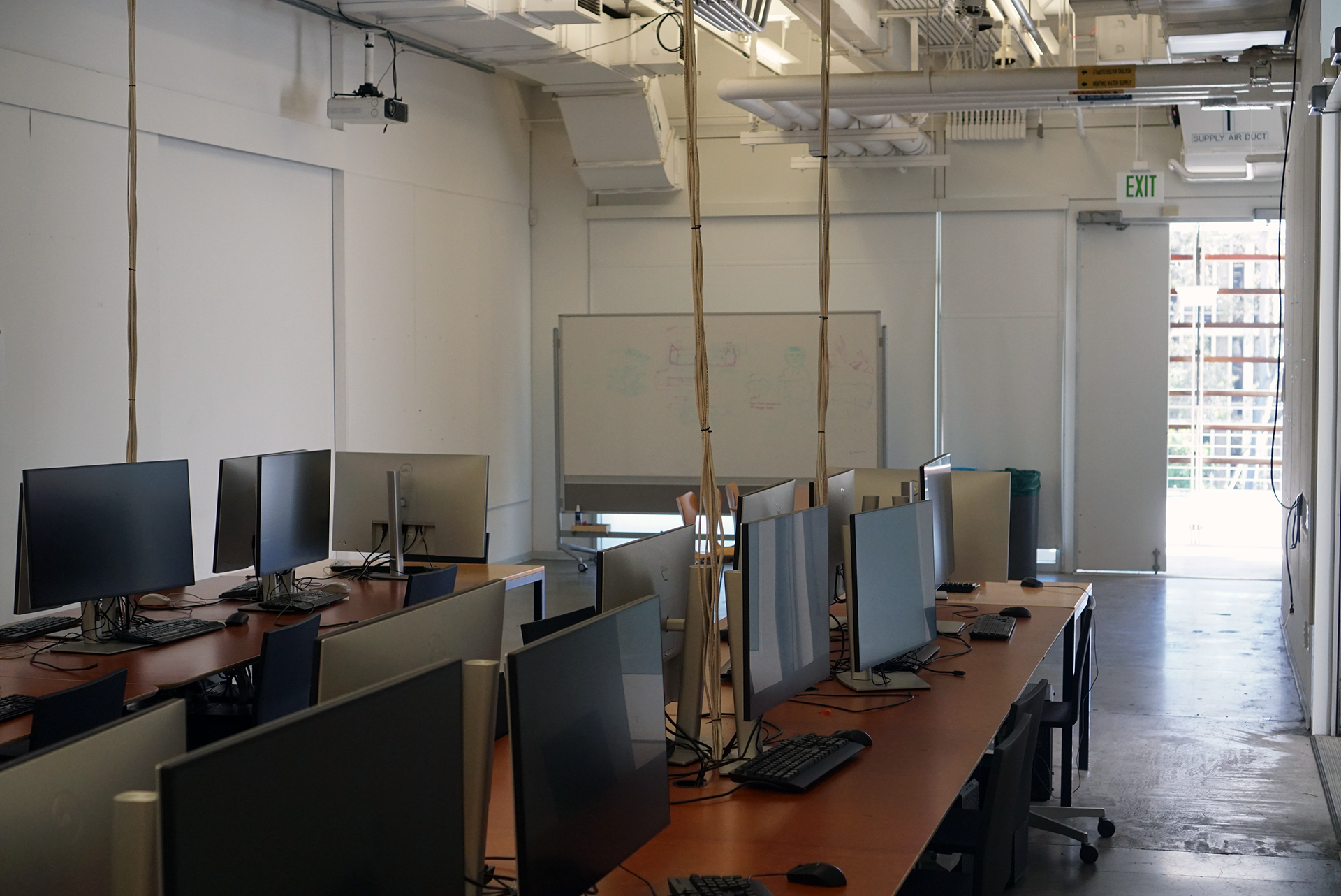 A view from the back of the Last Classroom computer lab, showing rows of wooden desks equipped with computer monitors and keyboards. In the background, there is a whiteboard and an open exit door letting in bright daylight.