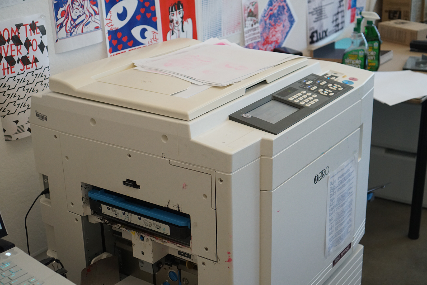 A large, off-white Risograph printer with a visible blue ink drum inside its open front panel, sitting in a workspace decorated with colorful graphic art posters on the wall behind it.